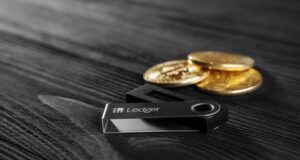 Is Ledger Preparing for an IPO? Here’s Why It Would Make Sense Ledger hardware wallet on wood background next to a stack of golden Bitcoin coins.